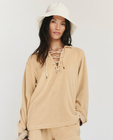 A woman wears THE GREAT. SU25 MICRO TERRY The Micro Terry Lace Up Tunic in Chamois and a cream bucket hat, posed against a neutral background.