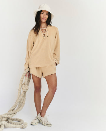 A woman wears THE GREAT. SU25 MICRO TERRY The Micro Terry Lace Up Tunic in chamois with matching shorts, white bucket hat, and sneakers, holding a thick rope against a plain light background.