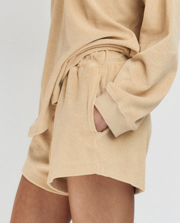 A person wears THE GREAT. SU25 MICRO TERRY The Micro Terry Swing Short in Chamois with a matching sweatshirt, one hand in the pocket; only their lower torso and upper legs are visible.