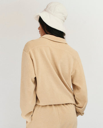 A person in THE GREAT. SU25 MICRO TERRY The Micro Terry Lace Up Tunic in Chamois and matching pants with a white bucket hat, facing away from the camera against a plain background.