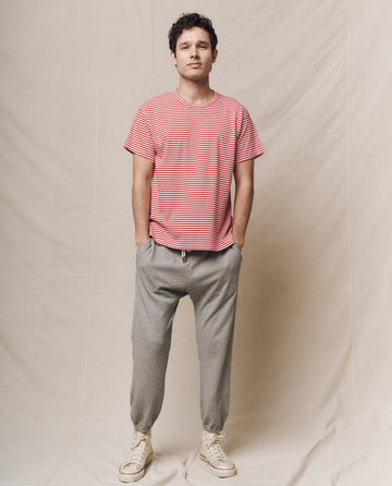 A person wearing THE GREAT.'s Men's Boxy Crew in Sun Cherry Stripe, gray sweatpants, and white sneakers stands against a beige backdrop.