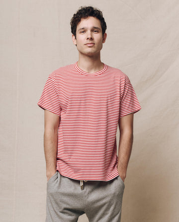 A person in a The Men's Boxy Crew shirt from THE GREAT. SP25 POLO LOUNGE MEN in Sun Cherry Stripe and gray pants stands with hands in pockets against a plain backdrop.