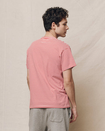 A person with curly hair wears The Men's Boxy Crew in Sun Cherry Stripe from THE GREAT. SP25 POLO LOUNGE MEN, paired with gray pants. They stand sideways against a plain background exuding casual elegance.