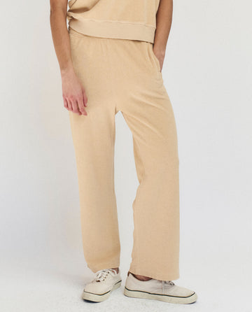 Model wears THE GREAT. SU25 MICRO TERRY The Micro Terry Victory Sweatpant in Chamois with white sneakers, standing against a plain light background. Only the lower half of the body is visible.