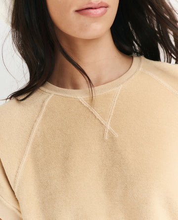 Close-up of a woman wearing THE GREAT. SU25 MICRO TERRY The Micro Terry Wedge Sweatshirt in Chamois, dark hair partially visible, shown from the shoulders up.