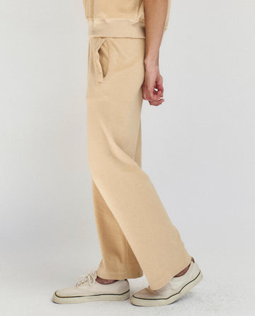 A person wearing THE GREAT. SU25 MICRO TERRY The Micro Terry Victory Sweatpant in Chamois and white sneakers stands against a plain light background.