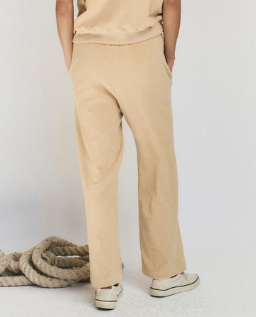 A person stands with hands in pockets, wearing THE GREAT. SU25 MICRO TERRY The Micro Terry Victory Sweatpant in Chamois and sneakers, facing away; a coiled rope rests on the floor nearby.