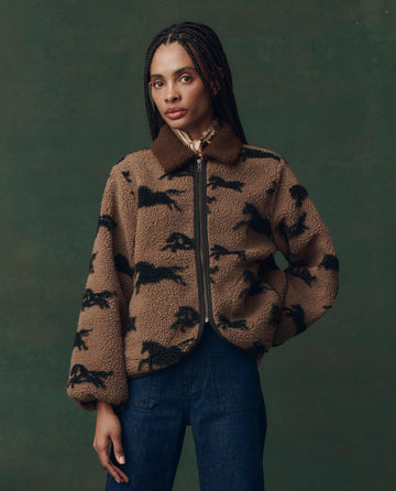 A woman with braids wears THE GREAT. F25 D2's The Pasture Jacket in Light Hickory, a plush brown zip-up with a black animal pattern, styled with dark blue jeans against a dark green background.