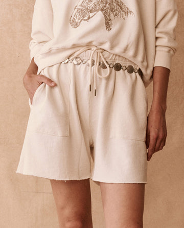 Person wearing The Patch Pocket Sweat Short in Washed White by THE GREAT. SU24 BIRKENSTOCK, featuring large front patch pockets and an elastic waistband with an ornate belt, along with a matching sweatshirt that features an animal design. Beige background.