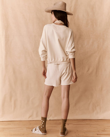A person wearing a hat, a light-colored long-sleeve top, matching 'The Patch Pocket Sweat Short' in Washed White from THE GREAT. SU24 BIRKENSTOCK, green socks with a design, and sandals stands with their back to the camera against a beige background.