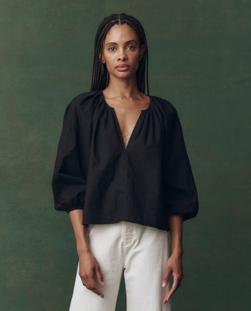 A person stands against a green background wearing THE GREAT. F25 D2’s The Chariot Top in textured black cotton with three-quarter sleeves and white pants, looking directly at the camera.