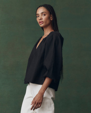 A woman poses against a green background, wearing THE GREAT. F25 D2’s The Chariot Top in black—a loose-fitting, textured cotton style with three-quarter sleeves—paired with white pants.