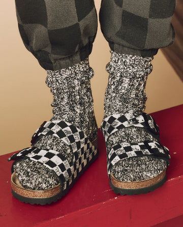 A person wearing The Hand Stamped BIRKENSTOCK Arizona in Black with Cream Check from THE GREAT. HOL 24 BIRKENSTOCK over black and white patterned socks is standing on a vibrant red surface.