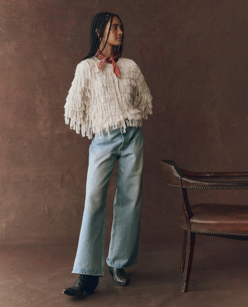 Indoors, a person wears THE GREAT. F25 D1’s The Fringe Cardigan in Natural, styled with light blue wide-leg jeans, black boots, and a red scarf, standing next to a brown leather chair against a brown background.