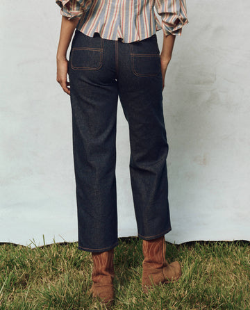 Person standing on grass, wearing a striped shirt, The Workwear Jean - Rinse by THE GREAT. PS25, paired with brown boots, facing away from the camera.