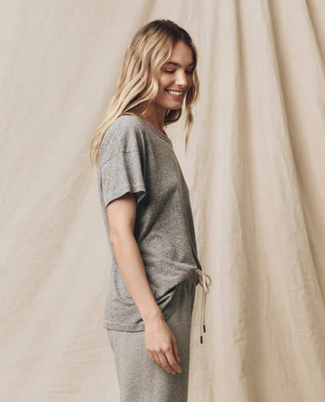 A woman with long blonde hair smiles while standing sideways, wearing a grey t-shirt and matching pants against a beige backdrop. Her Boxy Crew in Heather Grey by THE GREAT. CORE KNITS adds a touch of timeless style to the scene.