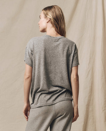 A person with long hair stands facing away, showcasing a loose-fitting vintage tee and gray pants against a beige backdrop. The Boxy Crew in Heather Grey from THE GREAT. CORE KNITS, made in the U.S.A., features flattering tweaks that set it apart.