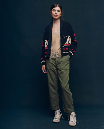 A person stands against a dark background, showcasing a vintage style featuring The Vintage Army Pant by THE GREAT. SP25 COLLECTORS. They wear a dark cardigan with red patterns, a beige top, and complete the look with classic white sneakers.