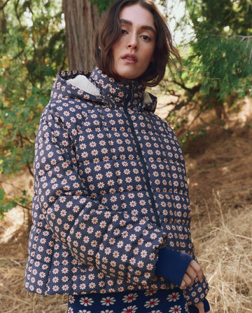A person wearing THE GREAT. F25 TGO's The Whistler Puffer in Mini Frosted Daisy with recycled insulation stands outdoors among trees and dry grass.
