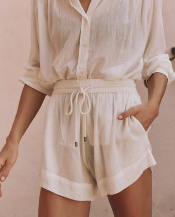 A person wears THE GREAT. SP25 SWIM's The Delta Short in White, styled high-waisted with a drawstring, paired with a semi-sheer white button-up shirt with rolled sleeves, standing against a light background.