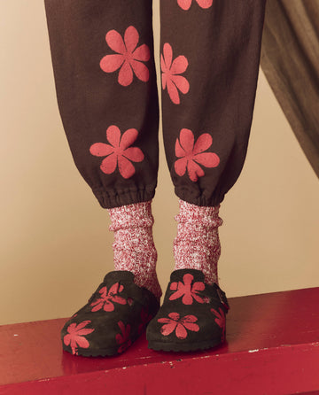 A person wearing The Hand Stamped Boston Birkenstock in Mocha with Hot Red Daisy from THE GREAT. HOL 24 BIRKENSTOCK, complemented by hand-stamped red and white speckled socks, stands confidently on a vibrant red surface.