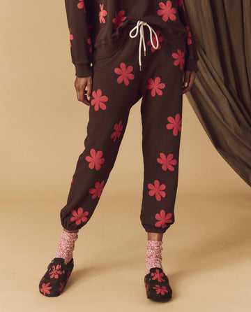 A person stands indoors on a beige floor, wearing brown joggers embellished with hot red daisy patterns and a matching top. Pink socks peek out from under their mocha Hand Stamped Boston Birkenstock slippers by THE GREAT. HOL 24 BIRKENSTOCK, creating a cozy look inspired by Boston's cool style.