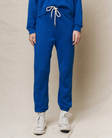 Dressed in bright cobalt Stadium Sweatpants with a white drawstring from THE GREAT. SP25 KNITS, paired with a matching top and sleek white high-top sneakers, someone stands against a neutral backdrop.