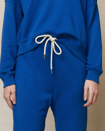 A person wearing The Stadium Sweatpant in Bright Cobalt from THE GREAT. SP25 KNITS, with a white drawstring at the waist, stands against a neutral background.