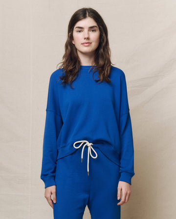 A person in The Teammate Sweatshirt in Bright Cobalt from THE GREAT. SP25 KNITS and matching pants stands against a beige background.