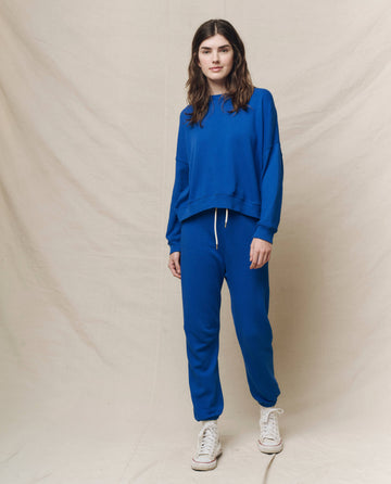 A person stands wearing THE GREAT. SP25 KNITS' The Teammate Sweatshirt in Bright Cobalt and joggers, paired with white sneakers, against a plain beige backdrop.