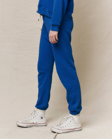 A person sports The Stadium Sweatpant in Bright Cobalt by THE GREAT. SP25 KNITS, pairing it with white high-top sneakers and striking a confident pose against a neutral background.