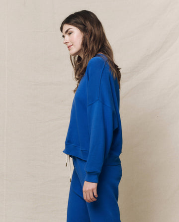 A woman in "The Teammate Sweatshirt" in Bright Cobalt by THE GREAT. SP25 KNITS, paired with pants, stands against a beige backdrop, facing left, epitomizing casual wear elegance.