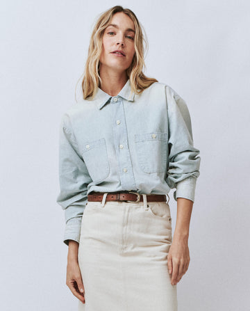 A person with long blonde hair is wearing The Road Shirt in Riverbed Wash by THE GREAT. FALL 24 COLLECTORS, paired with a beige skirt and standing against a plain background.