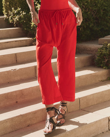 Person wearing The Reef Pant in Hot Red by THE GREAT. SP25 SWIM, featuring a comfortable fit, paired with black strappy sandals while standing on stone steps surrounded by greenery.