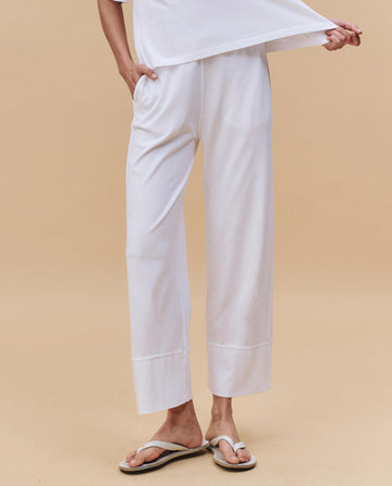 A person wears THE GREAT. PF25 HEAVY JERSEY KNITS The Jersey Win Pant in True White, paired with a white top and sandals, standing against a beige background—an effortlessly chic loungewear look.