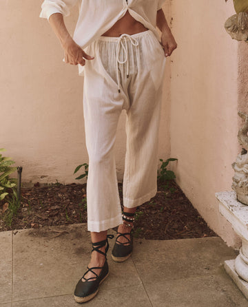 A person stands outdoors on a stone patio in the white Reef Pant by THE GREAT. SP25 SWIM and a matching top, paired with black lace-up espadrilles—a nod to sustainable women’s fashion. Face not visible.