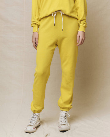 In vibrant athleisure, a person confidently stands on a beige surface, wearing THE GREAT. SP25 KNITS' The Stadium Sweatpant in Daffodil and white sneakers.