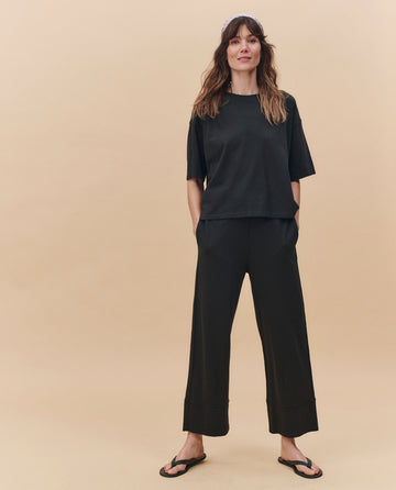 A woman stands against a beige background wearing THE GREAT. PF25 HEAVY JERSEY KNITS The Track Crew. in Almost Black, styled with wide-leg black pants and flip-flops, hands in her pockets and embracing an oversized silhouette.