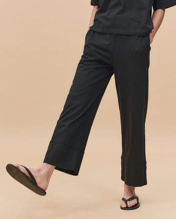 A person models The Jersey Win Pant in Almost Black by THE GREAT. PF25 HEAVY JERSEY KNITS, paired with a black top and flip-flops, standing on one leg against a beige background in effortlessly stylish loungewear.