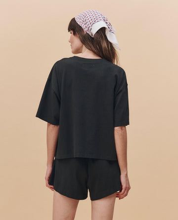 A woman faces away against a beige background, wearing THE GREAT. PF25 HEAVY JERSEY KNITS The Flutter Short in Almost Black, paired with an oversized black T-shirt and a white patterned headscarf.