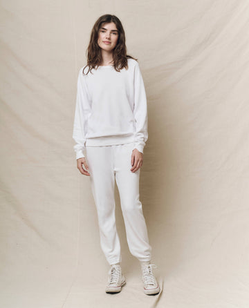 A person dressed in The Stadium Sweatpant. Solid by THE GREAT. HOL 24 KNITS, featuring an elasticated waist and paired with white sneakers, stands against a beige fabric backdrop.