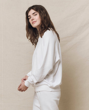 A person with long brown hair is wearing THE GREAT. HOL 24 KNITS' The College Sweatshirt in True White and pants, standing against a beige background.
