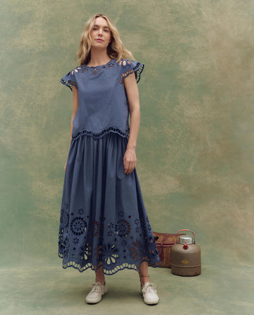 A woman stands before a green mottled backdrop, modeling THE GREAT. SU25’s Super Bloom Eyelet Top in Vintage Blue, paired with white sneakers. A vintage suitcase and jug rest on the floor behind her.