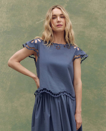 A woman with long blonde hair models THE GREAT. SU25’s The Super Bloom Eyelet Top in Vintage Blue, paired with a matching skirt against a green textured backdrop for an effortless, standout look.