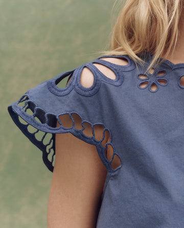 Close-up of a person wearing THE GREAT. SU25 The Super Bloom Eyelet Top in Vintage Blue, featuring cut-out lace details on the sleeves and neckline—a chic choice for any season against a plain background.