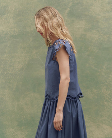 A woman with blonde hair stands in profile against a green textured background, wearing THE GREAT. SU25’s The Super Bloom Eyelet Top in Vintage Blue for an effortlessly stylish look.