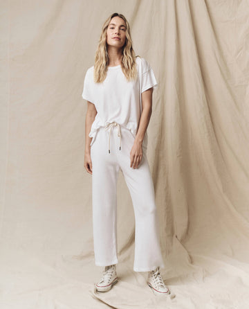A person is standing against a beige backdrop wearing The Boxy Crew in True White from THE GREAT. CORE KNITS, complemented by matching white drawstring pants and white sneakers.