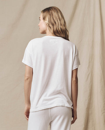 A person with long, light hair is standing with their back to the camera, wearing "The Boxy Crew. Solid -- True White" T-shirt by THE GREAT. CORE KNITS paired with white pants against a beige background.