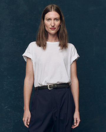 A woman with straight brown hair wears THE GREAT. CORE KNITS The Boxy Crew. Solid -- True White, a 100% cotton tee made in the USA, with dark pants, standing against a dark blue background facing the camera neutrally.