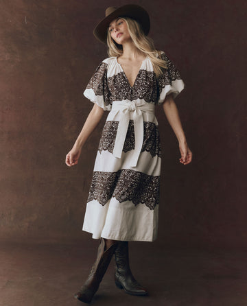 A woman wears THE GREAT. F25 D1 The Fairground Dress in cream with western lace print, styled with a wide belt, brown hat, and black boots as she stands against a dark background.